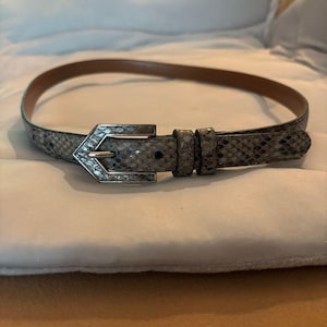 May include: A slim belt featuring a textured snakeskin pattern in shades of grey, brown, and blue. The belt includes a pointed, silver-toned metal buckle and two matching keepers. The interior of the belt strap is a smooth, solid tan-coloured material.