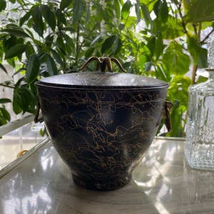 May include: A black ice bucket with a lid and gold-colored handles and accents. The bucket has a gold scribble pattern across the surface. The ice bucket is sitting on a white surface with green plants in the background.