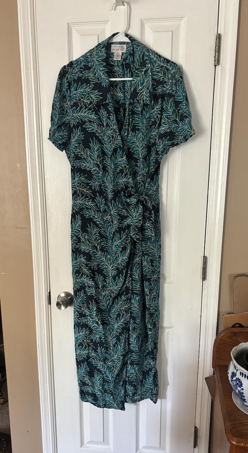 Puede incluir: Un vestido largo de color azul oscuro con un estampado de hojas verdes y blancas. El vestido tiene mangas cortas y un escote en V. La etiqueta de la marca dice "Papillon".