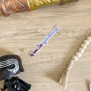 May include: A pink and blue lightsaber sticker with black details. The sticker is on a light brown wooden surface.