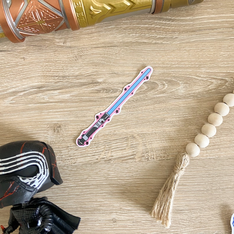 May include: A pink and blue lightsaber sticker with black details. The sticker is on a light brown wooden surface.