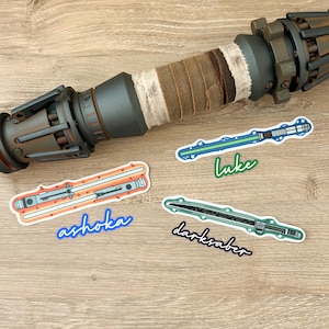 May include: Four Star Wars lightsaber stickers on a wooden surface. The stickers feature the lightsabers of Luke Skywalker, Ahsoka Tano, and the Darksaber. The stickers are printed on white paper with a glossy finish.