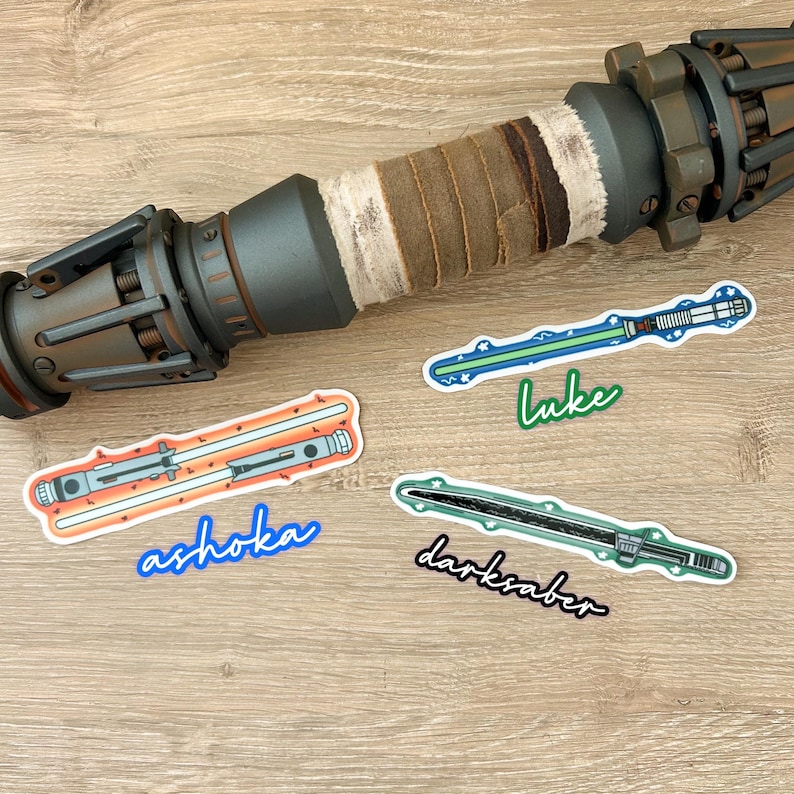 May include: Four Star Wars lightsaber stickers on a wooden surface. The stickers feature the lightsabers of Luke Skywalker, Ahsoka Tano, and the Darksaber. The stickers are printed on white paper with a glossy finish.