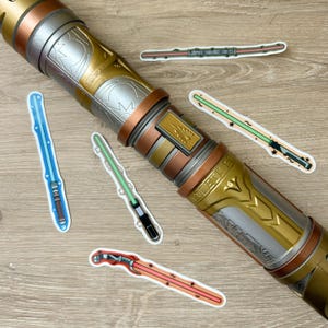 May include: A collection of five colourful lightsaber stickers on a wooden surface. The stickers feature different lightsaber designs, including a blue lightsaber, a green lightsaber, a red lightsaber, and a green lightsaber with a yellow hilt. The stickers are all in the shape of a lightsaber and have a shiny finish.