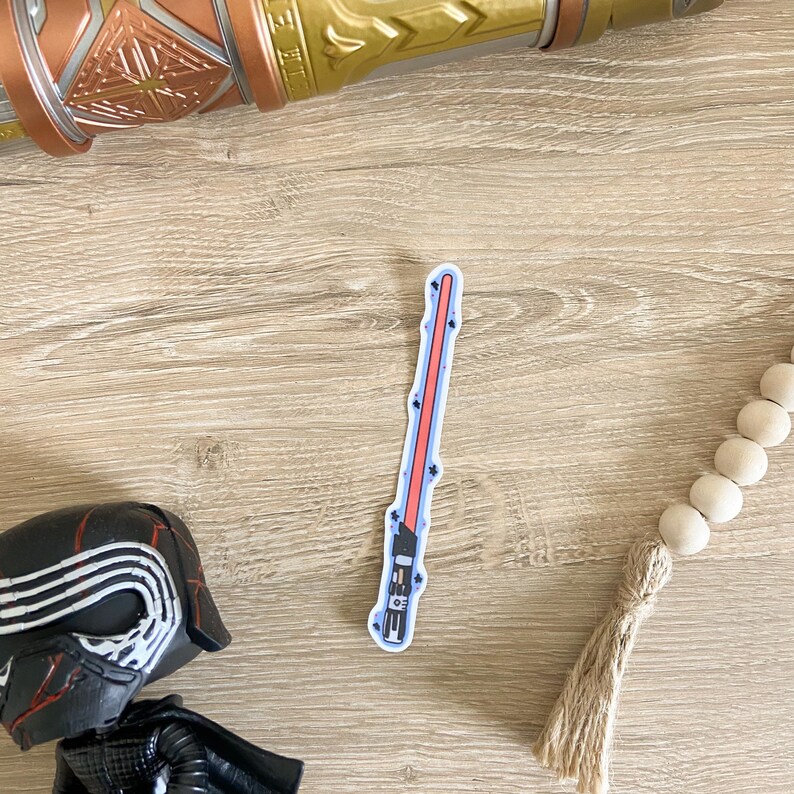 May include: A red lightsaber sticker with a black handle and blue accents. The sticker is on a light brown wooden surface.