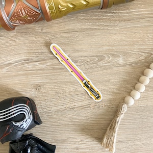 May include: A pink lightsaber sticker with a black handle and yellow accents. The sticker is on a wooden surface.
