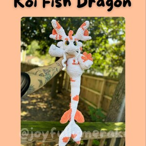 May include: A white and orange crocheted koi fish dragon plush toy. The toy has a long tail with orange and white stripes. The toy is being held by a person's hand.