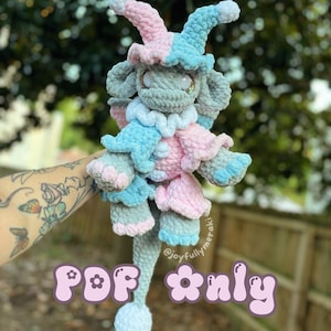 May include: A crocheted plush toy in the shape of a jester. The toy is gray with pink and blue accents. The toy has a long tail and is wearing a jester hat. The text "PDF only" is written in pink letters with a flower design.