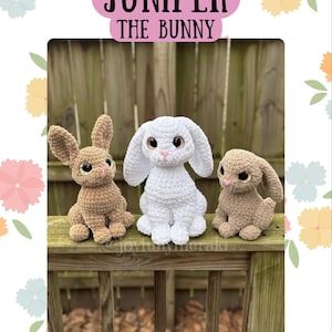 Może przedstawiać: Trzy szydełkowe figurki króliczków w odcieniach brązu i bieli są wystawione na drewnianej powierzchni. Tekst "JUNIPER THE BUNNY" znajduje się nad króliczkami. Króliczki mają duże uszy i ciemne oczy. Tło to drewniane ogrodzenie.