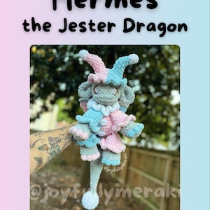 K&ouml;nnte beinhalten: Ein geh&auml;keltes Pl&uuml;schspielzeug-Drache mit einer Narrenm&uuml;tze in Rosa und Blau. Der Drache ist grau mit einem langen Schwanz und wird von einer Hand mit einem Tattoo gehalten. Das Bild enth&auml;lt den Text "Hermes the Jester Dragon" und den Social-Media-Handle "@joyfullymeraki".