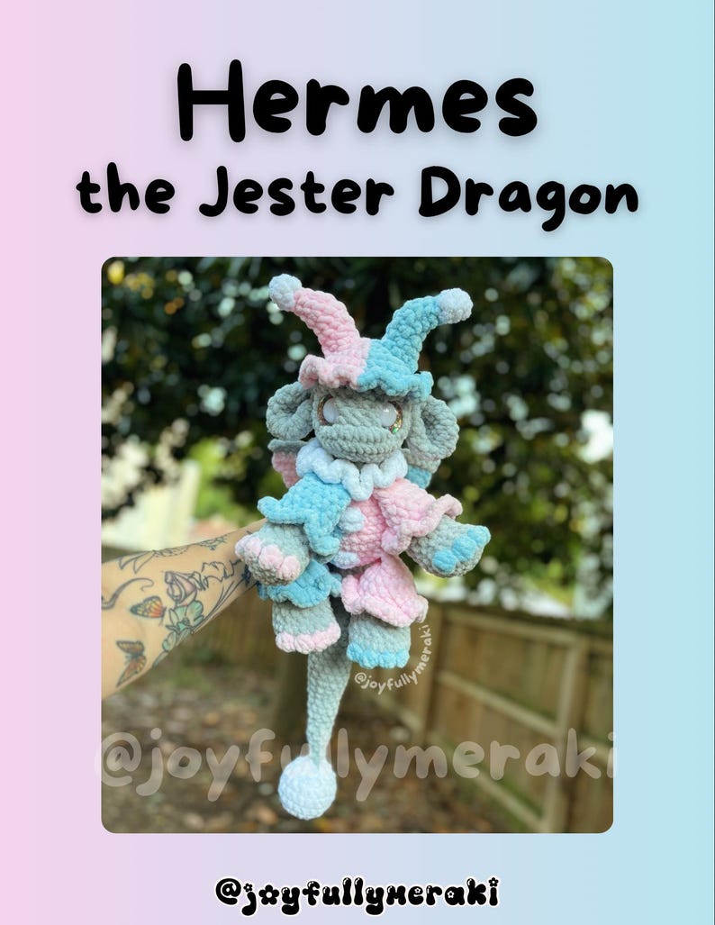 K&ouml;nnte beinhalten: Ein geh&auml;keltes Pl&uuml;schspielzeug-Drache mit einer Narrenm&uuml;tze in Rosa und Blau. Der Drache ist grau mit einem langen Schwanz und wird von einer Hand mit einem Tattoo gehalten. Das Bild enth&auml;lt den Text "Hermes the Jester Dragon" und den Social-Media-Handle "@joyfullymeraki".