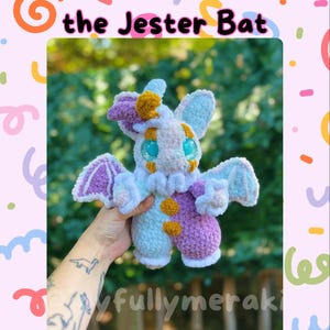 May include: A handmade plush bat toy in pastel colors. The bat is white, light blue, and purple with orange and yellow accents. The text "Baubo the Jester Bat" is above the toy. The background is a colorful design with the text "joyfullymeraki".