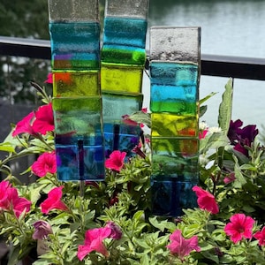 May include: Three rectangular glass garden stakes featuring stacked horizontal segments in shades of blue, green, and clear glass. Each stake is mounted on a thin metal rod and is positioned among vibrant pink petunia flowers in a dark planter.