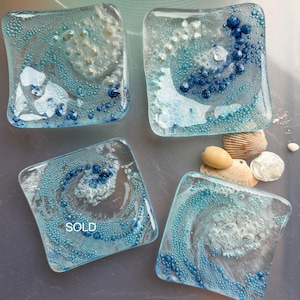 May include: Four square glass coasters with ocean-themed designs. Each coaster features a swirling pattern of blue and white, with embedded bubbles and small blue beads. The bottom left coaster has the word "SOLD" in white. Seashells are arranged near the coasters.