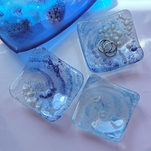 May include: A collection of handcrafted glass art pieces in shades of blue. The set includes a large, curved dish and several square dishes with swirling patterns and embedded bubbles. One square dish holds three silver rings.