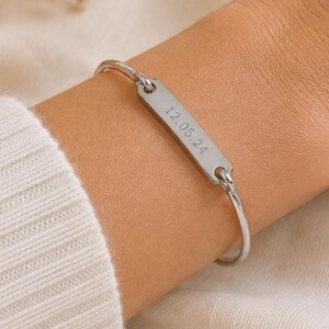 May include: A silver-coloured metal bangle bracelet featuring a central rectangular bar plate. The plate is engraved with the date 12.05.24 in a simple sans-serif font. The bracelet has a thin, polished wire band that connects to each end of the bar.