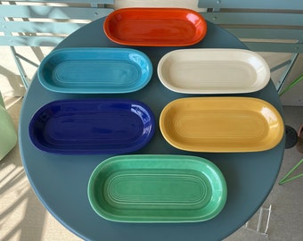 May include: Six colorful oval ceramic dishes in shades of blue, green, yellow, orange, white, and dark blue. The dishes are arranged on a blue table.