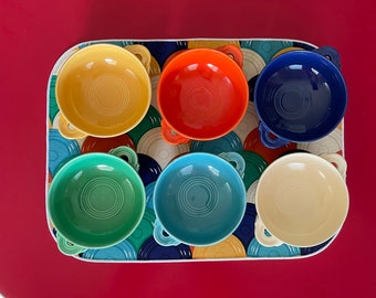 May include: Six colorful ceramic bowls with handles, arranged in a circle on a patterned mat. The bowls are yellow, orange, blue, green, turquoise, and cream.