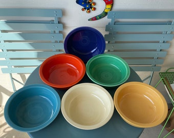 May include: A set of six brightly coloured ceramic bowls in shades of blue, red, green, cream, and yellow. The bowls are displayed on a blue metal table, with a decorative lizard and blue chairs in the background.