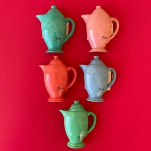 May include: Five colorful ceramic teapots in shades of green, coral, pink, and blue, arranged on a red background. Each teapot has a handle, spout, and lid, with the word "Fiesta" in a script font.