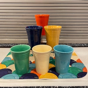 May include: A set of six ceramic tumblers in various colors including orange, dark blue, yellow, light green, cream, and light blue. Each tumbler features a tapered shape with horizontal ribbed detailing around the upper exterior surface.