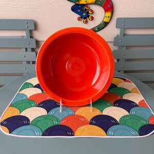 May include: A vibrant red Fiesta ware bowl sits on a colorful placemat with a repeating pattern of overlapping circles in various colors. A decorative metal lizard hangs in the background. The bowl is displayed on a clear plastic stand.