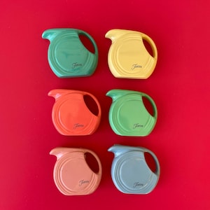 May include: Six colorful ceramic Fiesta ware pitchers in green, yellow, orange, light green, peach, and blue. Each pitcher has a handle and spout, with the "Fiesta" logo printed on the front. The pitchers are arranged on a red background.