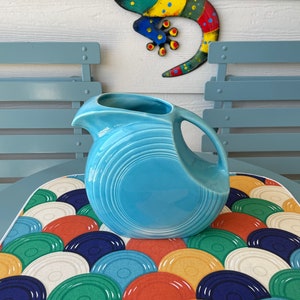 May include: A light blue ceramic pitcher with a rounded body and a wide mouth. The pitcher has a textured, swirling design and is sitting on a colorful, patterned placemat.