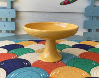 May include: A yellow ceramic pedestal bowl with a scalloped edge. The bowl is sitting on a colourful table runner with a pattern of overlapping circles.