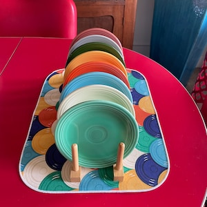May include: A set of colorful ceramic plates stacked on a wooden stand. The plates are arranged in a rainbow pattern, with the top plate being a light green color. The plates are sitting on a colorful placemat with a pattern of circles.