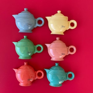 May include: Six colorful ceramic teapots in shades of blue, yellow, green, pink, orange, and teal. Each teapot has a handle, spout, and lid, with the word "Fiesta" printed on the front. The teapots are arranged on a red background.