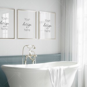 May include: Three gold framed pictures with the text "Your design here" hanging on a white wall above a white bathtub with a white towel draped over the edge. The bathtub is in a bathroom with a blue wall and a white window with white curtains.