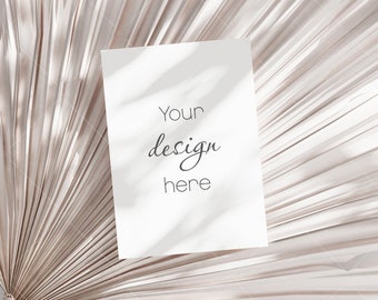 Puede incluir: Una tarjeta rectangular blanca con el texto "Your design here" en un estilo manuscrito, apoyada sobre una hoja de palma beige texturizada. La imagen está iluminada con luz natural suave, creando sombras.