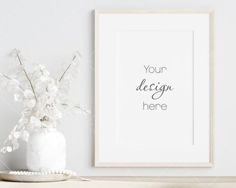 May include: A white frame with a white mat and a white background. The text "Your design here" is written in black cursive script. The frame is hanging on a white wall. A white vase with white branches is in the foreground.