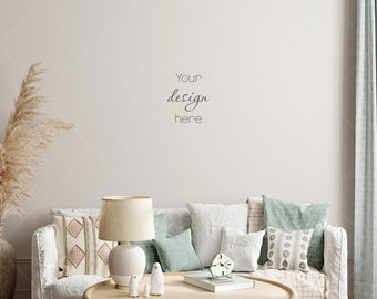 May include: A white couch with a variety of pillows in shades of beige, white, and teal. A table lamp with a white shade and a white base sits on a wooden coffee table in front of the couch. The coffee table also has a book, a clock, and three small white figurines. The wall behind the couch is white and has the text "Your design here" written on it.