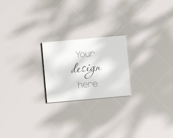 Puede incluir: Una tarjeta blanca con el texto "Your design here" escrito en letra cursiva negra. La tarjeta está sobre una superficie blanca con sombras de una planta frondosa.