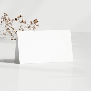 May include: A blank white folded card stands upright on a reflective white surface. A sprig of dried, beige-colored flowers rests near the card. The background is a light, textured white.