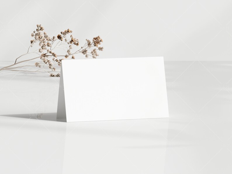 May include: A blank white folded card stands upright on a reflective white surface. A sprig of dried, beige-colored flowers rests near the card. The background is a light, textured white.