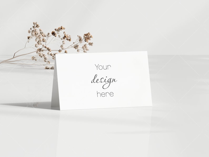 May include: A white folded card with the text "Your design here" in a handwritten style. Dried, light brown flowers are placed to the left of the card. The card is on a white, reflective surface.