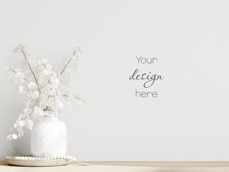 Op de afbeelding: Witte keramische vaas met een boeket gedroogde witte bloemen op een houten tafel tegen een witte muur. De tekst "Your design here" staat op de muur.