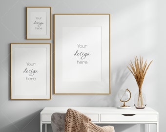 Op de afbeelding: Drie gouden fotolijsten met witte passe-partouts hangen aan een lichtgrijze muur boven een wit bureau met een vaas met gedroogd tarwe en een globe op een witte standaard. De foto's hebben de tekst "Your design here" in elk.