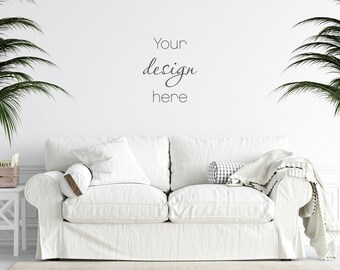 May include: A white sofa with a linen slipcover, adorned with pillows and a blanket. The wall behind the sofa has the text "Your design here". Palm fronds frame the scene, suggesting a tropical or coastal theme.