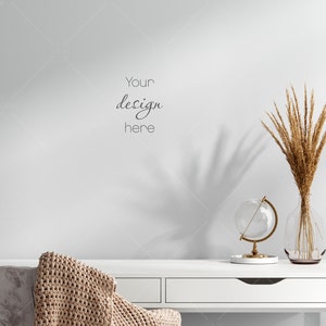 Puede incluir: Escritorio blanco con dos cajones, una manta de punto beige drapeada sobre una silla, un globo terráqueo de vidrio sobre un soporte dorado y un jarrón con espigas de trigo secas. El texto "Your design here" está escrito en la pared.