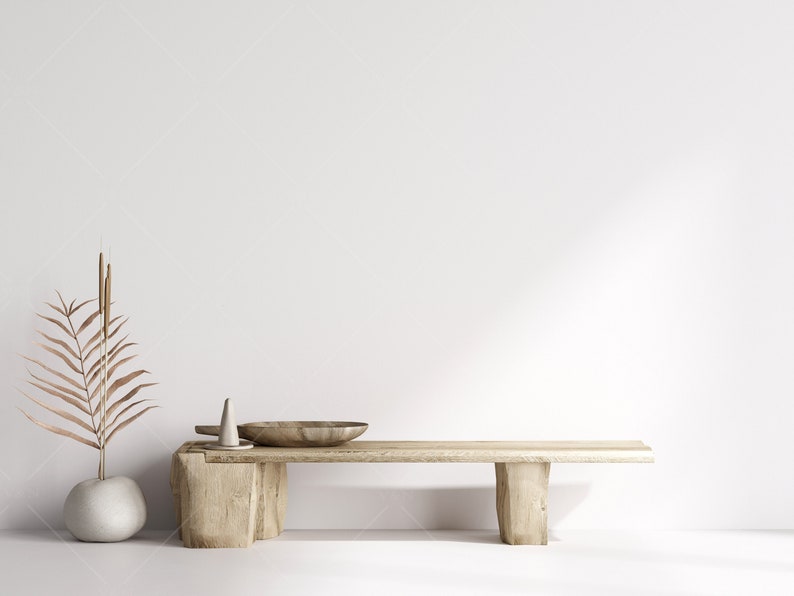 Pu&ograve; includere: Una panca in legno con una finitura naturale si trova di fronte a un muro bianco. Un piccolo vaso bianco con foglie di palma marroni secche si trova sul pavimento a sinistra della panca. Una ciotola di legno e un piccolo cono di ceramica bianca si trovano sopra la panca.