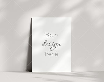 Puede incluir: Un papel rectangular blanco apoyado contra una pared blanca. El texto "Your design here" está escrito en una fuente cursiva. La imagen está iluminada con luz natural, proyectando sombras.
