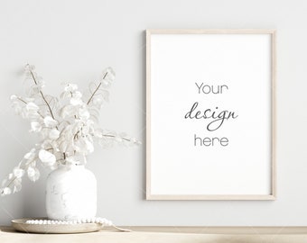 May include: A white vase with dried white flowers sits on a wooden table next to a light wood picture frame with the text "Your design here".