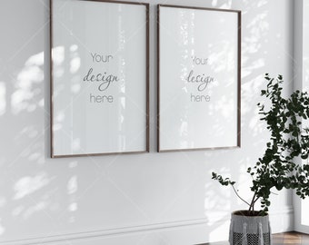 May include: Two framed art prints with brown wooden frames are mounted on a white wall. The prints have white backgrounds with the text "Your design here" in a script font. A potted plant with green leaves sits on a wooden floor.