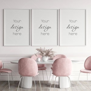 Op de afbeelding: Drie witte fotolijsten met de tekst "Your design here" hangen aan een witte muur boven een witte tafel met vier roze stoelen. Een vaas met gedroogde bloemen staat op de tafel.