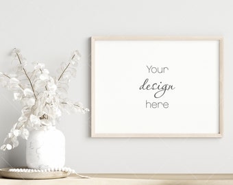 May include: A white vase with dried white flowers sits on a wooden table in front of a white wall with a light wood frame around a blank white poster that says "Your design here".