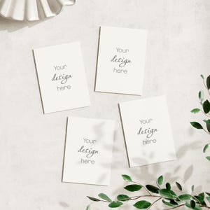 May include: Four blank white cards with the text "Your design here" on each card. The cards are arranged in a square shape on a white surface with green leaves and shadows.